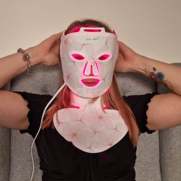 Woman adjusting red light therapy LED face mask and neck pad for full-coverage treatment.