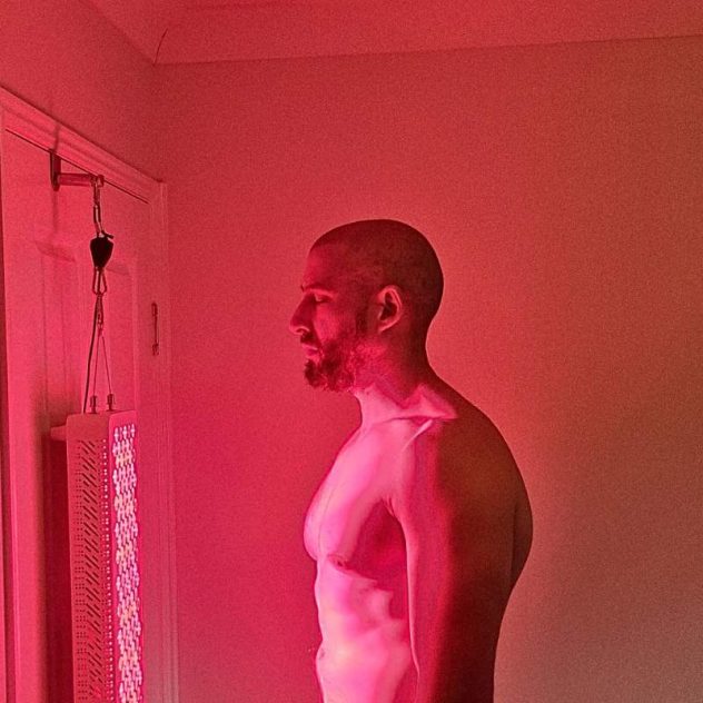 Man standing shirtless in front of a red light therapy panel for full-body treatment, skin rejuvenation, and muscle recovery.